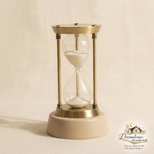 Peut inclure: Un sablier avec un cadre en laiton brossé et des ampoules en verre transparent contenant du sable blanc. Le sablier est monté sur une base circulaire beige, solide et lisse. Le logo Dreamhome decoratioUS avec le texte Turn your house into the home of your dreams est visible dans le coin inférieur droit.