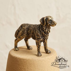 Puede incluir: Una pequeña figura metálica de color bronce de un perro de pie en posición de perfil. La escultura presenta un pelaje texturizado detallado, orejas puntiagudas y una expresión concentrada, capturando la apariencia de un perro retriever.