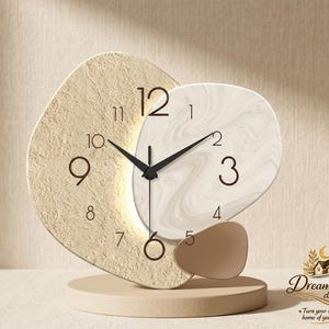 Puede incluir: Un reloj de mesa con un marco beige texturizado asimétrico y una esfera blanca con patrón de mármol. Cuenta con manecillas negras para las horas y minutos, números negros y una pequeña pieza decorativa marrón sobre una base circular.