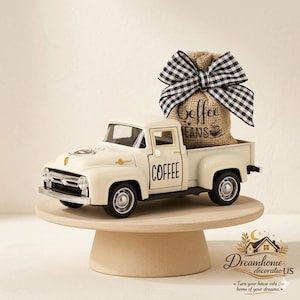 Puede incluir: Un modelo de camioneta vintage de color crema con la palabra COFFEE impresa en la puerta. En la caja de la camioneta hay un saco de arpillera con la etiqueta COFFEE BEANS, rematado con un gran lazo de tela de cuadros negros y blancos.