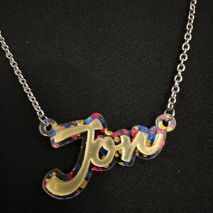 Peut inclure: Un collier en or avec un pendentif transparent qui dit "Joru" dans une police de caractères stylisée. Le pendentif est rempli de paillettes colorées.