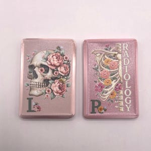 Pode incluir: Dois marcadores retangulares cor-de-rosa com acabamento brilhante. O marcador esquerdo apresenta um crânio humano adornado com flores cor-de-rosa e a letra L. O direito mostra uma caixa torácica humana com flores coloridas, a palavra RADIOLOGY e a letra R.