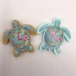 May include: Two turtle-shaped ornaments featuring a detailed shell pattern with tropical flowers, coral, and sea life. One turtle has a gold-toned border with the letter L, while the other has a light blue border with the letter R in pink.