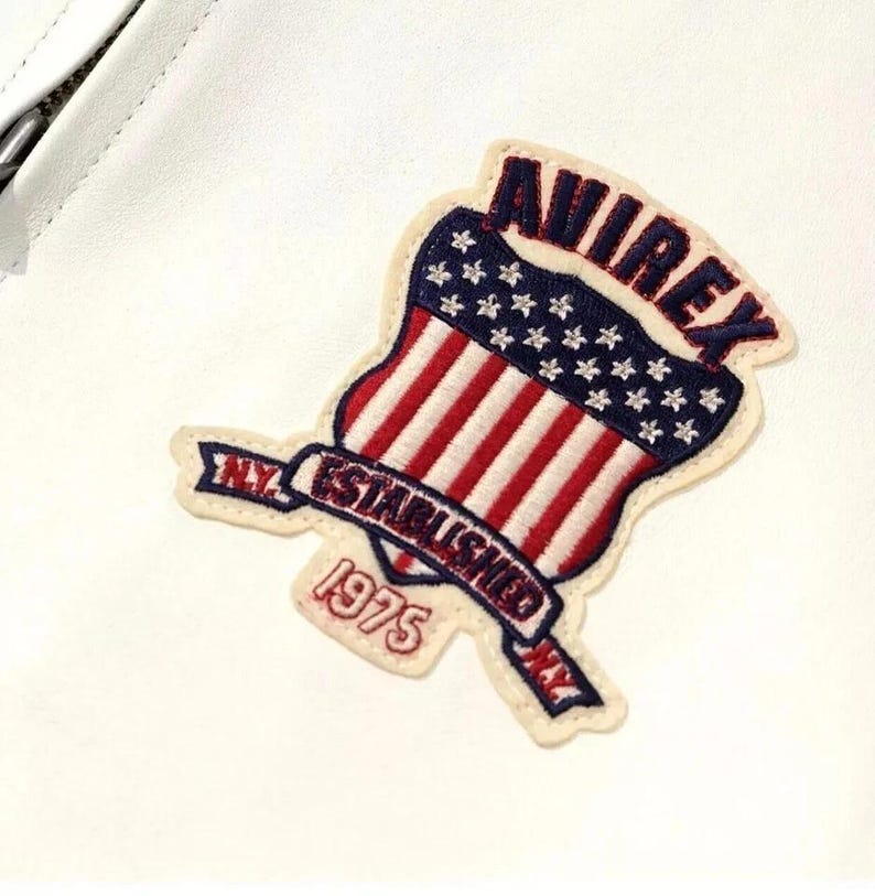May include: Embroidered patch featuring a shield design with red and white stripes and a blue field of stars. The patch reads "AVIREX" and "ESTABLISHED 1975" with "N.Y." on the banner. The patch is on a white background.