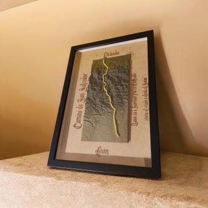 Puede incluir: Un mapa en relieve 3D enmarcado de la ruta del Camino de San Salvador, con un paisaje verde texturizado y una línea de sendero amarilla brillante. El mapa incluye el texto Oviedo, León y Camino de San Salvador en una fuente gótica negra.