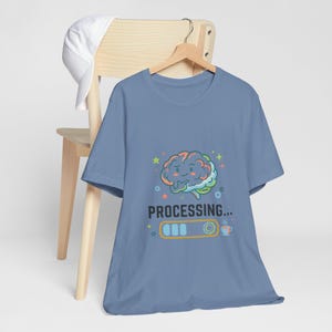 Può includere: Una t-shirt blu polvere con la grafica di un cervello a cartone animato con un'espressione pensierosa. Sotto il cervello, il testo "PROCESSING..." appare sopra una barra di caricamento e l'icona di una tazzina di caffè, circondato da stelle.