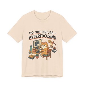 May include: Beige t-shirt featuring a cartoon cat working at a desk with a computer, desk lamp, and headphones. The text "DO NOT DISTURB - HYPERFOCUSING" is printed above the cat. The cat is orange and white, and the desk has various items.