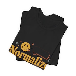 May include: A black t-shirt featuring a graphic print with the word Normalize in large yellow letters. The design includes a yellow smiley face with a small red heart, a speech bubble outline, and several small star shapes in red and blue colours.
