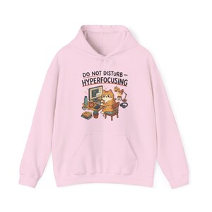 May include: A light pink hoodie with a front pocket and a drawstring hood. The front features a cartoon illustration of a cat working on a computer with the text "DO NOT DISTURB - HYPERFOCUSING" above the cat.