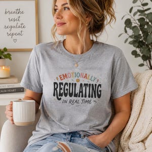 Può includere: Una maglietta grigia con la scritta "Emotionally Regulating in Real Time" in un carattere colorato e giocoso. La maglietta presenta anche una grafica con una faccina sorridente. L'immagine include una stampa incorniciata con le parole "breathe, reset, regulate, repeat."