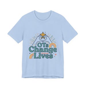 Peut inclure: Un t-shirt bleu clair à manches courtes avec une impression graphique portant le texte OTs Change Lives. Le motif représente deux mains formant un cœur autour d'une étoile jaune, entourées de petites icônes de cuillère, crayon et pince à linge.