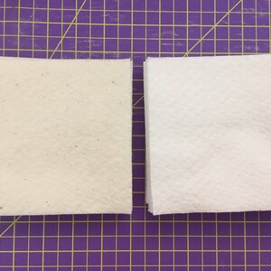 May include: Two stacks of white fabric squares, one with a slightly off-white color and a speckled texture, on a purple grid cutting mat.