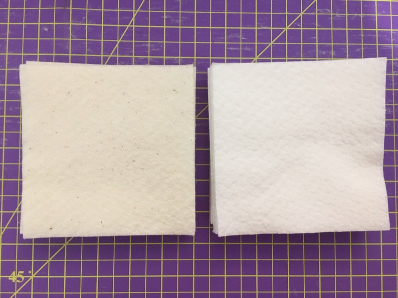 May include: Two stacks of white fabric squares, one with a slightly off-white color and a speckled texture, on a purple grid cutting mat.
