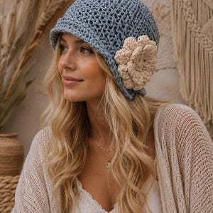 May include: A light blue crochet hat with a beige flower accent. The hat has a brim and is made of a textured yarn. The flower is attached to the side of the hat. The hat is a stylish accessory.