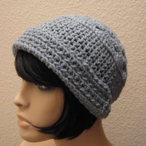May include: A grey crocheted beanie hat. The hat is made of a textured yarn and has a folded brim. The hat is shown on a mannequin head with black hair.