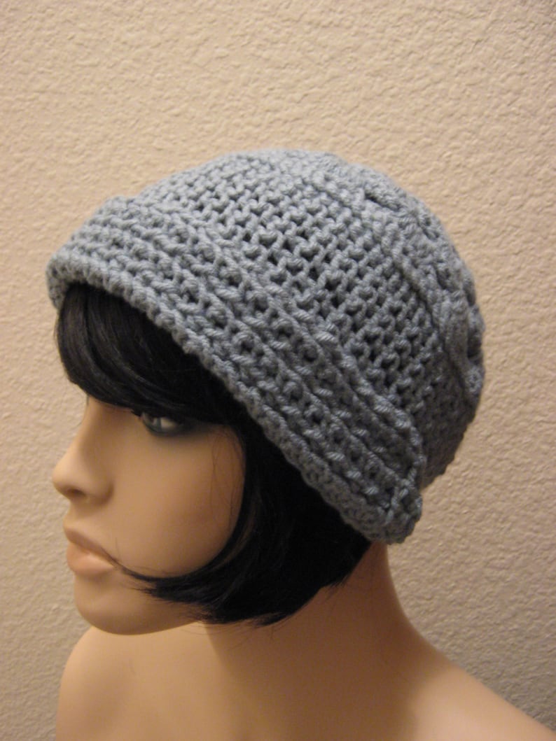 May include: A grey crocheted beanie hat. The hat is made of a textured yarn and has a folded brim. The hat is shown on a mannequin head with black hair.
