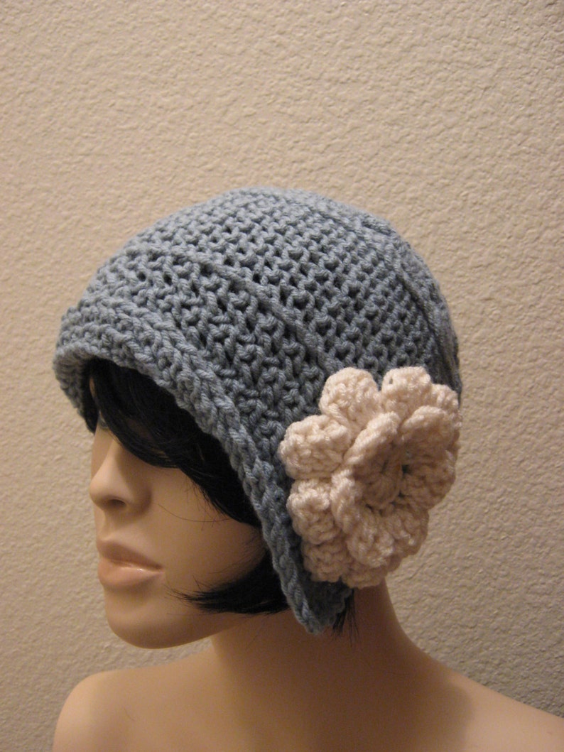 May include: A pale blue crocheted hat with a cream-coloured flower. The hat features a rounded crown and a small brim. A cream flower is attached to the side. The hat is made from a soft, textured yarn.