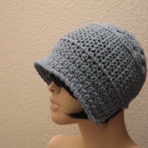 May include: A crocheted hat in a soft, muted blue-grey colour. The hat features a small, structured brim and a close-knit texture, offering a classic and versatile style. The hat is shown on a mannequin.