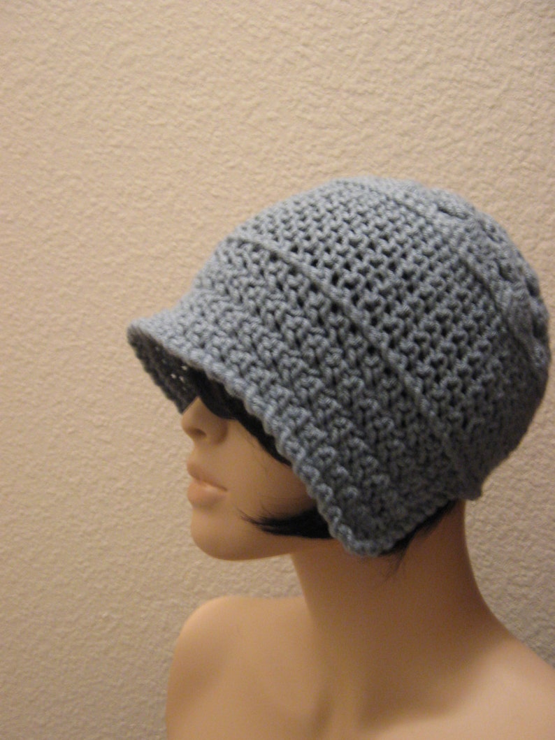 May include: A crocheted hat in a soft, muted blue-grey colour. The hat features a small, structured brim and a close-knit texture, offering a classic and versatile style. The hat is shown on a mannequin.