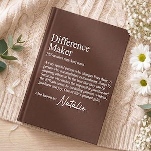 Puede incluir: Un libro de tapa dura de color marrón oscuro con texto en blanco que define el término Difference Maker, incluyendo una pronunciación fonética y un párrafo descriptivo, con el nombre Natalie impreso en la parte inferior como entrada también conocida como.