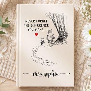 May include: A cream-coloured hardcover book featuring a black ink illustration of Winnie the Pooh and Piglet walking near trees. The text reads, "NEVER FORGET THE DIFFERENCE YOU MAKE..." with a small red heart and the name "mrs.sophia" below.