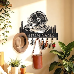 Könnte beinhalten: Ein schwarzer Wand-Schlüsselhalter aus Metall mit einem DJ-Motiv, Kopfhörern und einem Plattenspieler. Das Design umfasst eine horizontale Leiste mit dem Text CUSTOM NAME und mehrere Haken, an denen derzeit Schlüssel und eine braune Tasche hängen.