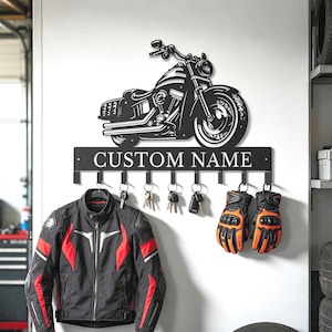 Può includere: Un portachiavi da parete in metallo nero con una silhouette di motocicletta sopra una barra orizzontale con il testo CUSTOM NAME. Il supporto include otto ganci per appendere oggetti, con diverse chiavi e un paio di guanti neri e arancioni.