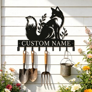 May include: A black metal wall-mounted rack featuring a silhouette of a fox sitting among foliage. The rack includes a horizontal bar with the text CUSTOM NAME and eight hooks, currently holding three garden tools and a small metal bucket.