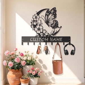 Può includere: Un portachiavi da parete in metallo nero con un intaglio decorativo a forma di mezzaluna, fiori e farfalla. La barra orizzontale presenta la scritta CUSTOM NAME e otto ganci per appendere oggetti come chiavi, una borsa e delle cuffie.