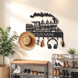 May include: A black metal wall-mounted hook rack shaped like a steam locomotive with smoke billowing from the top. The design features the text YOUR NAME and includes multiple hooks for hanging items like a straw hat, keys, and headphones.