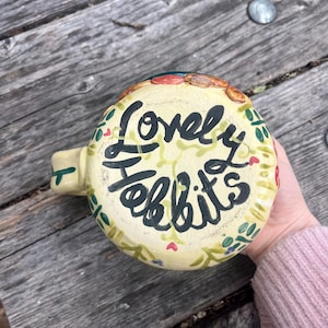 May include: A cream-colored ceramic mug featuring the hand-painted black text "Lovely Hobbits" on the base. The exterior is decorated with a painted pattern of green vines, small leaves, and tiny red hearts encircling the central lettering.