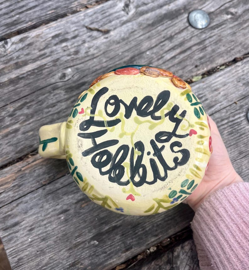 May include: A cream-colored ceramic mug featuring the hand-painted black text "Lovely Hobbits" on the base. The exterior is decorated with a painted pattern of green vines, small leaves, and tiny red hearts encircling the central lettering.