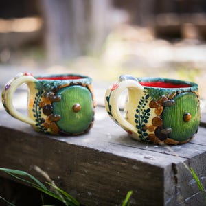 May include: Two ceramic mugs featuring a cream-colored exterior with a round green door design, brown stone-like accents, and a bright red interior. Each mug has a curved handle decorated with small painted floral details and a central knob.