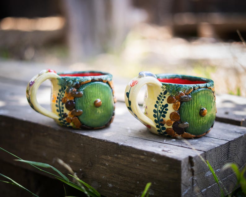May include: Two ceramic mugs featuring a cream-colored exterior with a round green door design, brown stone-like accents, and a bright red interior. Each mug has a curved handle decorated with small painted floral details and a central knob.