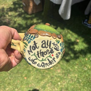 May include: A ceramic mug with a cream-colored base featuring the black hand-painted text Not all those who wander are lost. The top of the mug is decorated with a raised green textured dome surrounded by small brown stones and green leaf motifs.