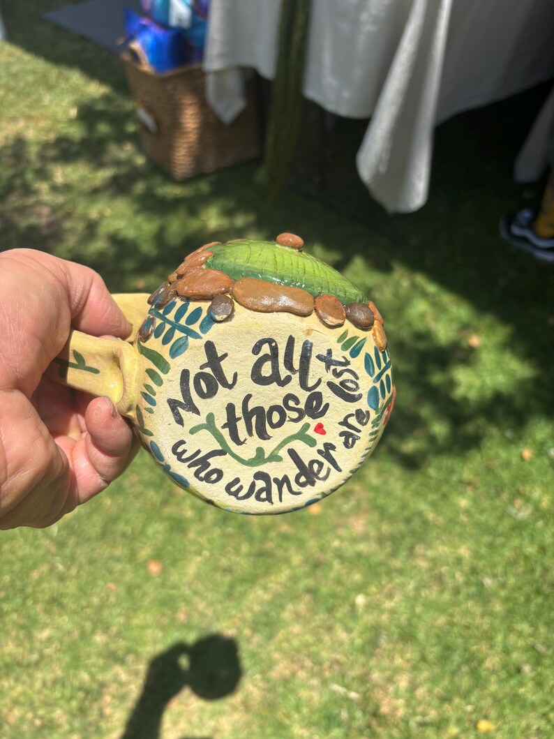 May include: A ceramic mug with a cream-colored base featuring the black hand-painted text Not all those who wander are lost. The top of the mug is decorated with a raised green textured dome surrounded by small brown stones and green leaf motifs.