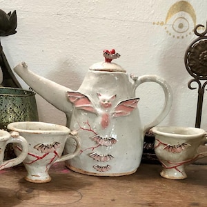 May include: A white ceramic teapot with a pink bat design and red accents. The teapot has a spout and handle and is decorated with a red heart on the lid. There are three matching teacups with pink bat designs and red accents.