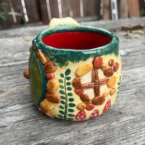 May include: A ceramic mug with a cream-colored exterior featuring raised, textured clay decorations including a brown cross-shaped window, small red and brown pebbles, green leaf patterns, and red mushroom shapes with white spots. The interior is bright red.