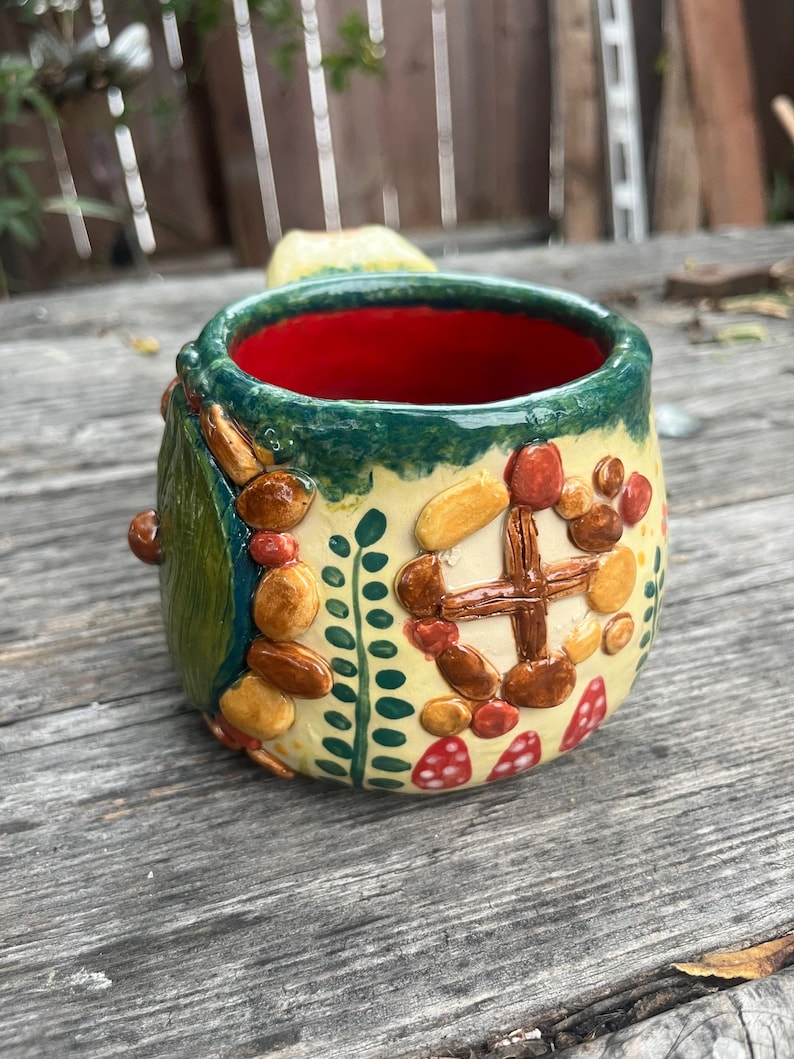 May include: A ceramic mug with a cream-colored exterior featuring raised, textured clay decorations including a brown cross-shaped window, small red and brown pebbles, green leaf patterns, and red mushroom shapes with white spots. The interior is bright red.