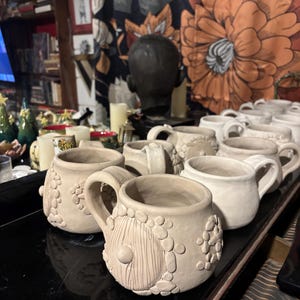May include: A collection of unglazed, light beige ceramic mugs featuring a round, textured body with a prominent circular door relief and small pebble-like clay accents. Each mug has a sturdy handle and a wide opening, arranged in rows.