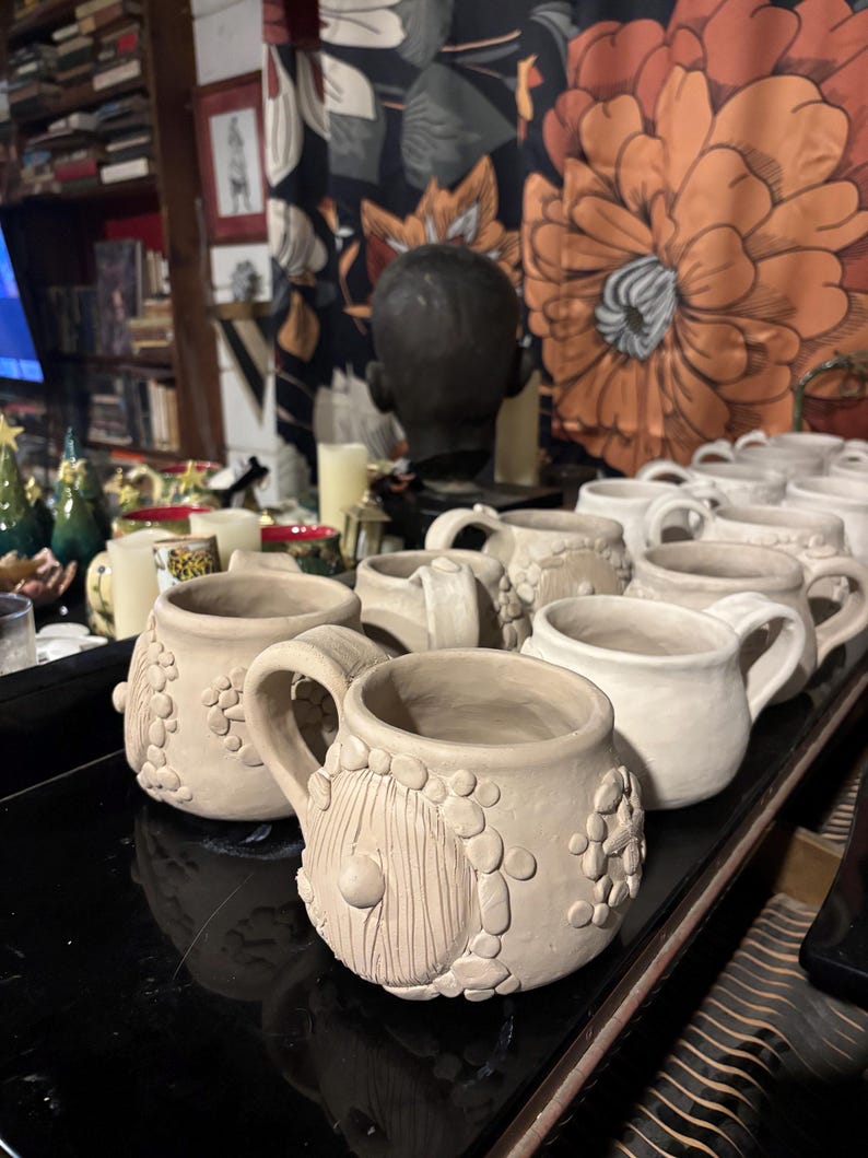 May include: A collection of unglazed, light beige ceramic mugs featuring a round, textured body with a prominent circular door relief and small pebble-like clay accents. Each mug has a sturdy handle and a wide opening, arranged in rows.