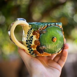 May include: A ceramic mug featuring a sculpted green circular door with a central knob, surrounded by brown stone-like textures and green foliage patterns. The mug has a cream-colored handle and a glossy finish with detailed relief work.