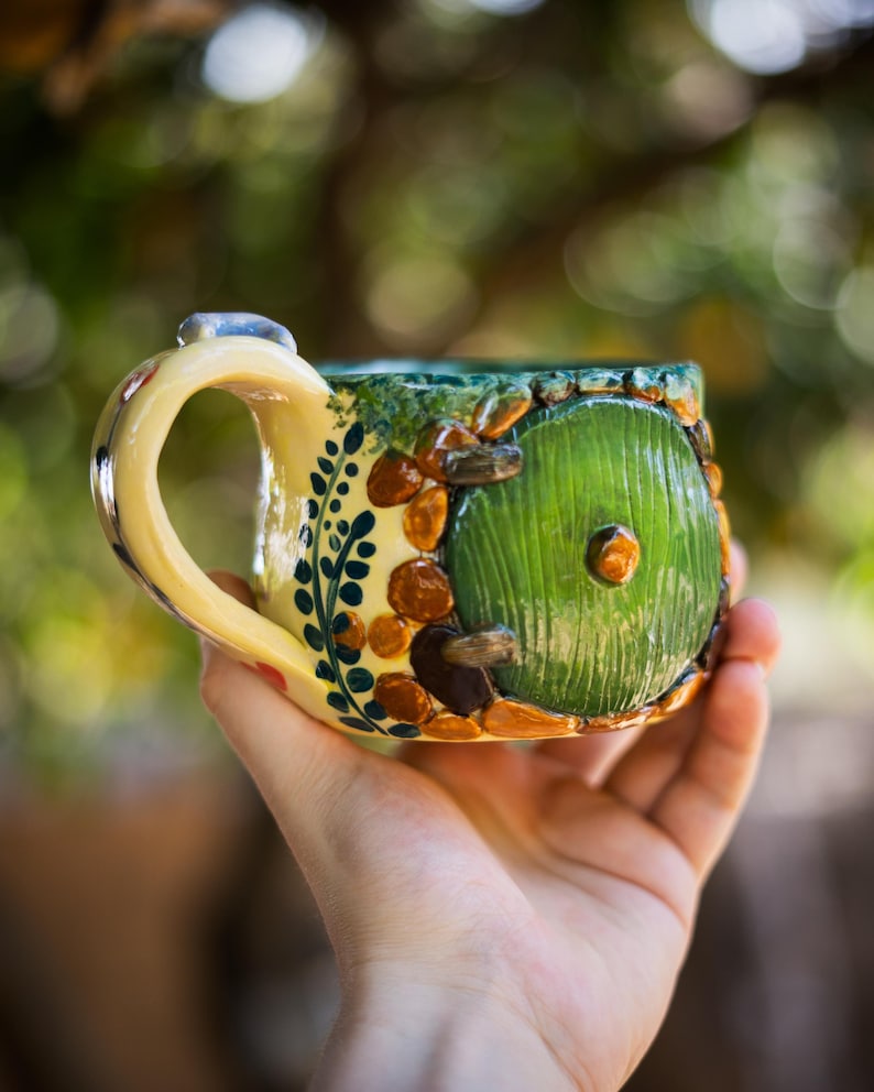 May include: A ceramic mug featuring a sculpted green circular door with a central knob, surrounded by brown stone-like textures and green foliage patterns. The mug has a cream-colored handle and a glossy finish with detailed relief work.