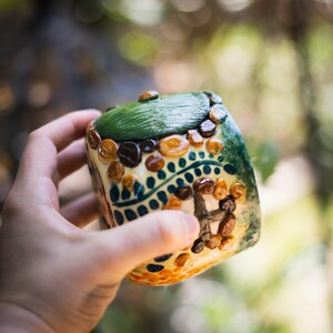 May include: A small, cylindrical ceramic container featuring a hand-painted design of a house with a green roof, brown stone-like accents, and blue decorative patterns. The surface has a glossy finish with raised textures depicting a door and walls.