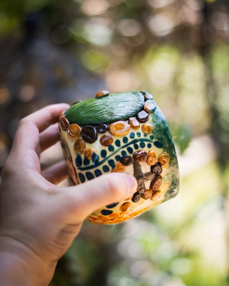 May include: A small, cylindrical ceramic container featuring a hand-painted design of a house with a green roof, brown stone-like accents, and blue decorative patterns. The surface has a glossy finish with raised textures depicting a door and walls.