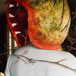 K&ouml;nnte beinhalten: Eine Person tr&auml;gt eine gro&szlig;e, rote, gr&uuml;ne und braune Pappmach&eacute;-Maske in Form einer fleischfressenden Pflanze mit einem gro&szlig;en, offenen Mund und wei&szlig;en Z&auml;hnen. Die Maske ist an einem wei&szlig;en T-Shirt befestigt.