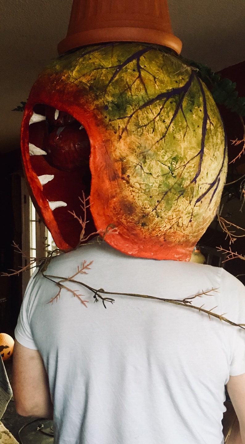 K&ouml;nnte beinhalten: Eine Person tr&auml;gt eine gro&szlig;e, rote, gr&uuml;ne und braune Pappmach&eacute;-Maske in Form einer fleischfressenden Pflanze mit einem gro&szlig;en, offenen Mund und wei&szlig;en Z&auml;hnen. Die Maske ist an einem wei&szlig;en T-Shirt befestigt.
