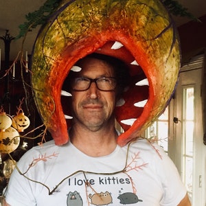 K&ouml;nnte beinhalten: Eine Person tr&auml;gt ein wei&szlig;es T-Shirt mit dem Text "Ich liebe K&auml;tzchen" und einer Grafik mit 12 Katzen. Die Person tr&auml;gt ein gro&szlig;es, rotes, pflanzenartiges Kost&uuml;m mit einem gro&szlig;en Mund und Z&auml;hnen. Das Kost&uuml;m ist aus Papiermach&eacute; gefertigt und mit einem Terrakotta-Topf gekr&ouml;nt.