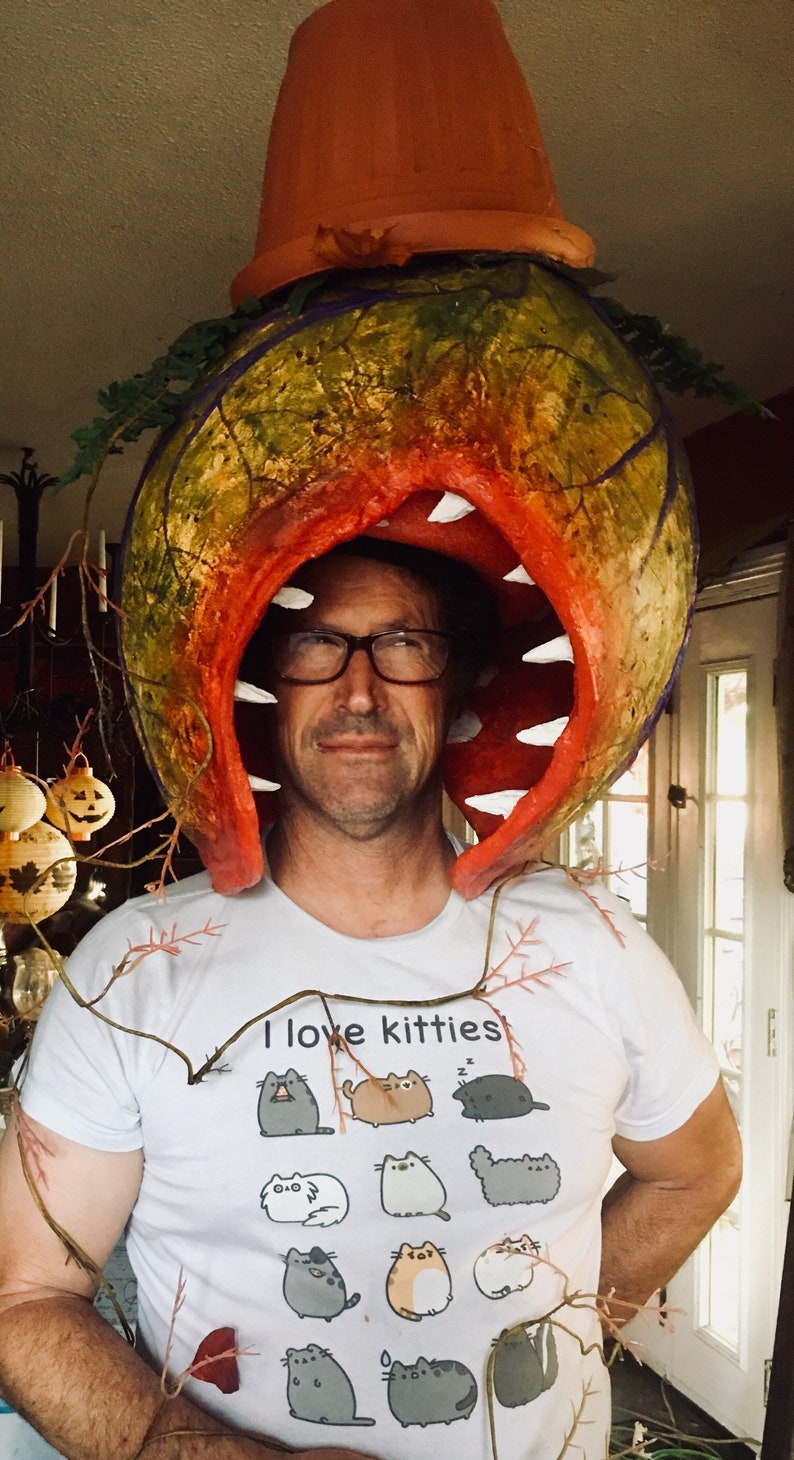 K&ouml;nnte beinhalten: Eine Person tr&auml;gt ein wei&szlig;es T-Shirt mit dem Text "Ich liebe K&auml;tzchen" und einer Grafik mit 12 Katzen. Die Person tr&auml;gt ein gro&szlig;es, rotes, pflanzenartiges Kost&uuml;m mit einem gro&szlig;en Mund und Z&auml;hnen. Das Kost&uuml;m ist aus Papiermach&eacute; gefertigt und mit einem Terrakotta-Topf gekr&ouml;nt.