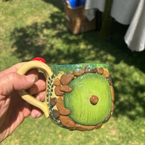 May include: A ceramic mug featuring a sculpted green circular door design surrounded by brown stone-like textures. The mug has a light tan handle and a green rim, with small blue leaf patterns painted along the side near the handle attachment.